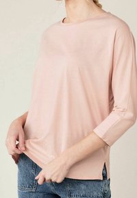 Helles rosa, langärmliges Oberteil aus weichem Stoff; mit rundem Ausschnitt, seitlichen Schlitzen und lockerer Passform. Kombiniert mit Jeans.