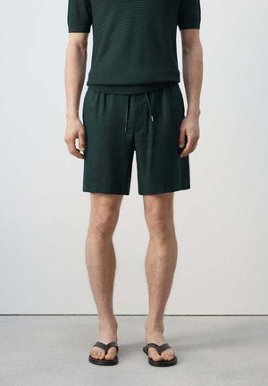 Hombre con camisa negra texturizada de manga corta, pantalones cortos negros con cordón y chanclas negras, de pie frente a un fondo blanco liso.