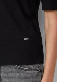 Zwarte gebreide trui met korte mouwen, een soepele textuur en een klein zilveren logo-label op de onderkant links. Gecombineerd met grijze denim.