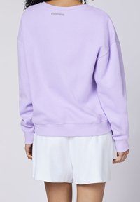 Sudadera de color púrpura claro con una tela texturizada, corte relajado y puños acanalados. Presenta un sutil logo en la parte trasera del cuello. Combinada con pantalones cortos blancos.