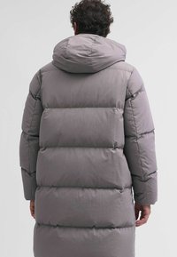 Grijze pufferjas met een capuchon, met een gestikte ontwerp en een verlengde vorm. De stof heeft een zachte textuur en zichtbare stiksels.