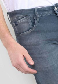 Vaqueros de denim gris oscuro con una textura suave, que cuentan con un bolsillo lateral, costuras visibles y un cierre de botón en la cintura.