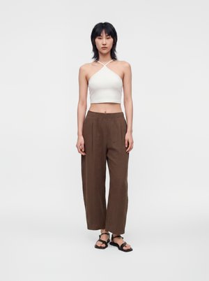 Osoba stoi, ubrana w biały crop top typu halter, luźne brązowe spodnie i czarne sandały na jednolitym białym tle.