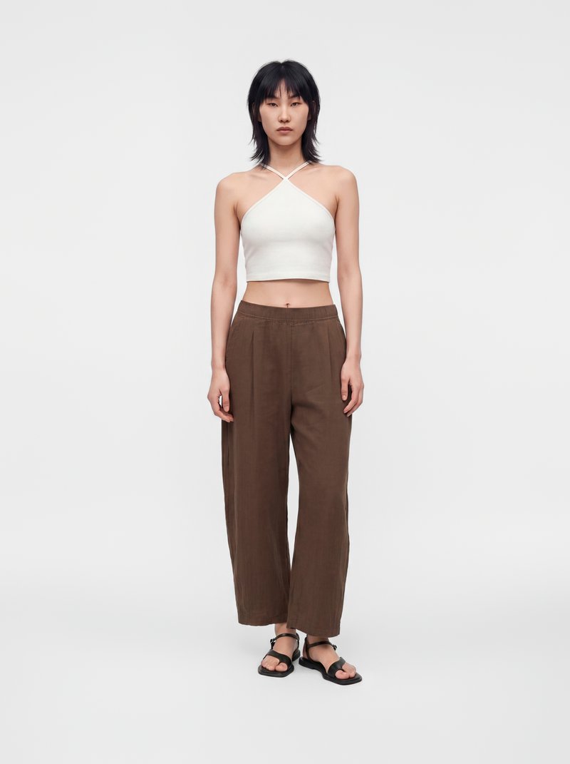 Personne debout, portant un crop top blanc à encolure américaine, un pantalon marron ample et des sandales noires sur un fond blanc uni.