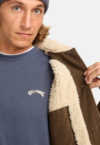Brown jacket with a cream sherpa lining, worn open over a blue sweater featuring a white logo. The jacket has a textured, sturdy material.