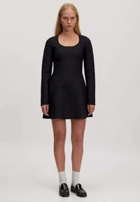 Schwarzes langärmliges Kleid mit quadratischem Ausschnitt, tailliertem Oberteil, ausgestelltem Rock und glatter Textur. Kombiniert mit schwarzen Slippern und weißen Socken.