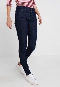Person som bär mörkblå slim fit jeans, vita sneakers och en långärmad tröja med röd- och vitrandigt, står mot en enfärgad bakgrund.