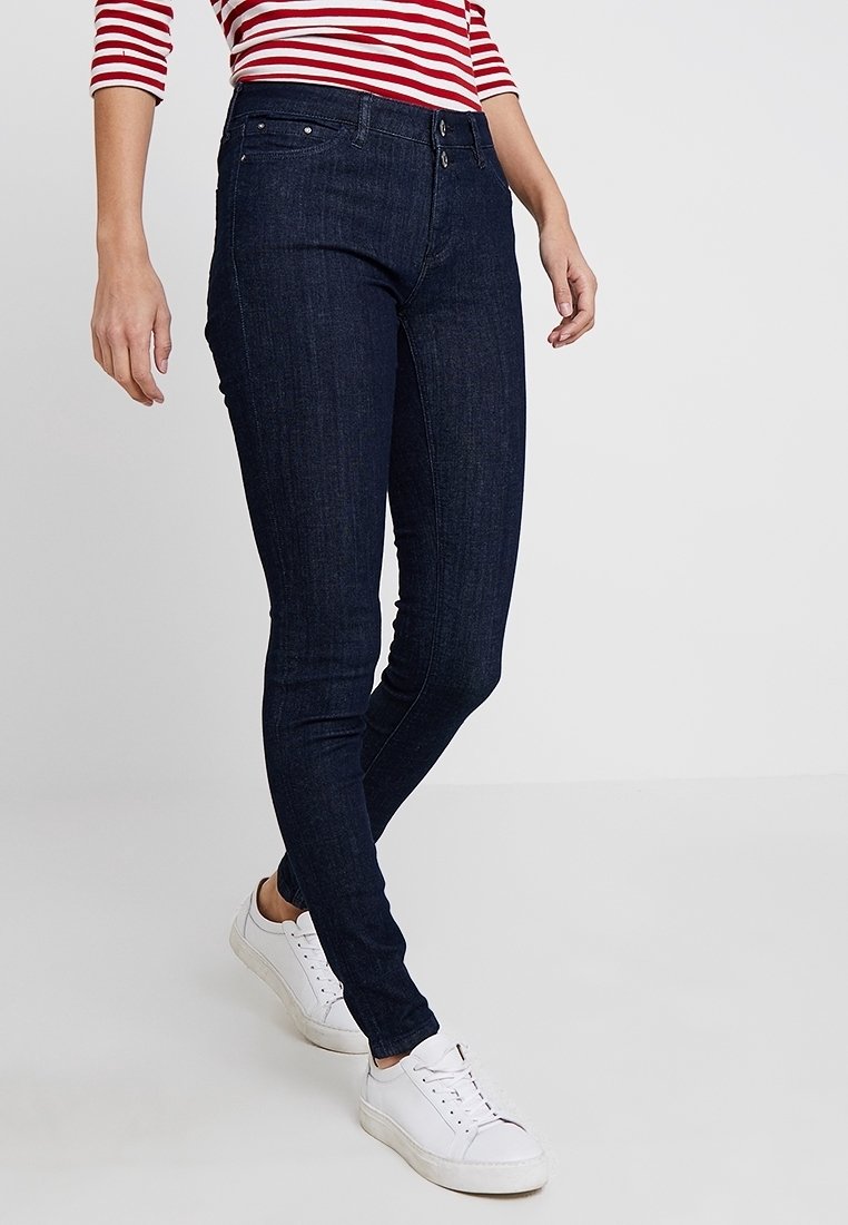 Person som bär mörkblå slim fit jeans, vita sneakers och en långärmad tröja med röd- och vitrandigt, står mot en enfärgad bakgrund.
