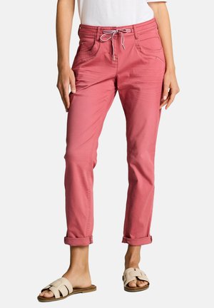 Kobieta w podwiniętych różowych spodniach z przodu z kieszeniami i białym troczkiem, do tego beżowe sandały wsuwane.