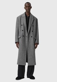 Divrindas pelēks herringbone mētelis ar lielām melnām pogām, sānu kabatām un brīvu piegriezumu, pārī ar melniem biksēm un apaviem.