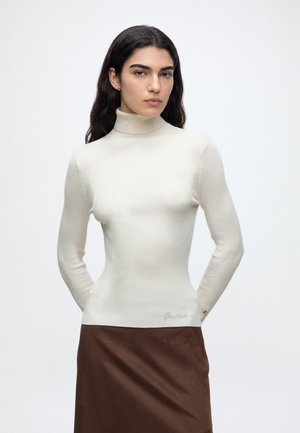 Femme aux longs cheveux foncés portant un pull à col roulé crème et une jupe marron, debout les mains derrière le dos.