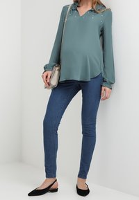 Blusa teal con scollatura a V, decorata con dettagli in perle, abbinata a jeans skinny blu scuro e ballerine nere. Borsa a tracolla appoggiata sulla spalla.