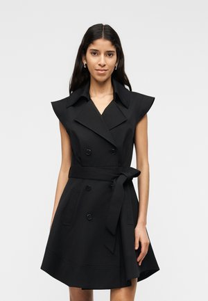 Femme portant une robe noire sans manches à double boutonnage avec un large col et une ceinture nouée, debout devant un fond clair uni.