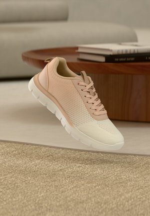 Lys beige atletiksneaker med hvid sål, svævende over et tekstureret beige tæppe i et moderne stueopslag.