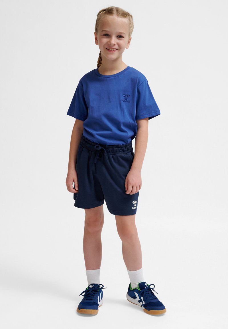 Blå kortärmad t-shirt med logotyp, tillsammans med mörkblå shorts med elastisk midja. Vita ankelstrumpor och marinblå sportskor med gröna accenter.