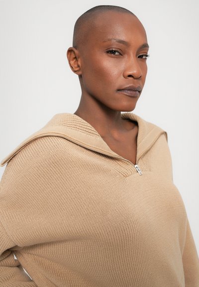 Beige Strickpullover mit halber Reißverschlusskragen, strukturiertem Design und lockerer Passform. Merkmale sind ein weiter Ausschnitt und Rippdetails entlang der Kanten.