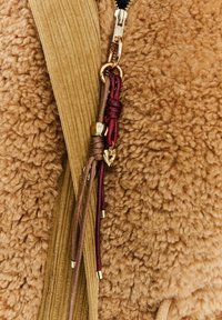 Detailní záběr na béžovou fleecovou bundu se zlatým zipem, béžovým kordovým lemováním a dekorativními hnědými a burgundskými uzlovými provázky se zlatými akcenty.