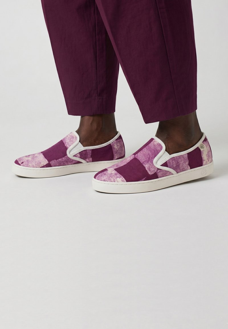 Pieds portant des baskets slip-on avec un motif patchwork violet et rose et des semelles blanches, assortis à un pantalon violet raccourci.