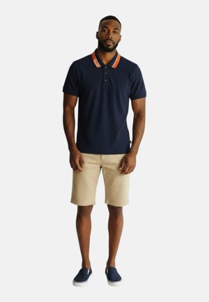 Uomo in piedi che indossa una polo blu navy con colletto arancione, pantaloncini beige e scarpe blu navy slip-on su sfondo bianco uniforme.