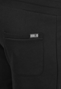 Zwarte joggerbroeken met ribbelachtige tailleband, voorzien van een klein achterzakje en een label met de tekst "ISOLID." Zachte stofstructuur, casual ontwerp.