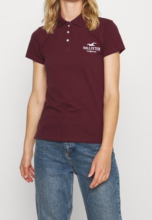 Kobieta ubrana w bordową koszulkę polo Hollister i niebieskie dżinsy, stojąca na tle jednolitego białego tła.