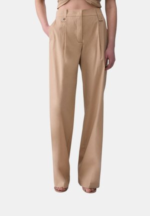 Pantaloni beige a vita alta e gamba larga con pieghe frontali e passanti per cintura con bottoni, indossati con le mani nelle tasche e sandali con punta aperta.
