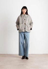 Giacca trapuntata grigia e crema a quadri con ampie tasche frontali, chiusura a bottoni e colletto, abbinata a pantaloni in denim blu a gamba larga e scarpe nere.