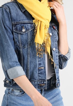 Personne portant une veste en denim retroussée, un jean bleu clair, une chemise blanche et une écharpe jaune à franges, avec une main près du visage.