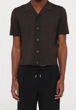 Mann trägt ein kurzärmeliges, schwarzes Hemd mit Knöpfen und Kragen sowie schwarze Jogginghosen mit Kordelzug, steht vor einem einfarbigen Hintergrund.