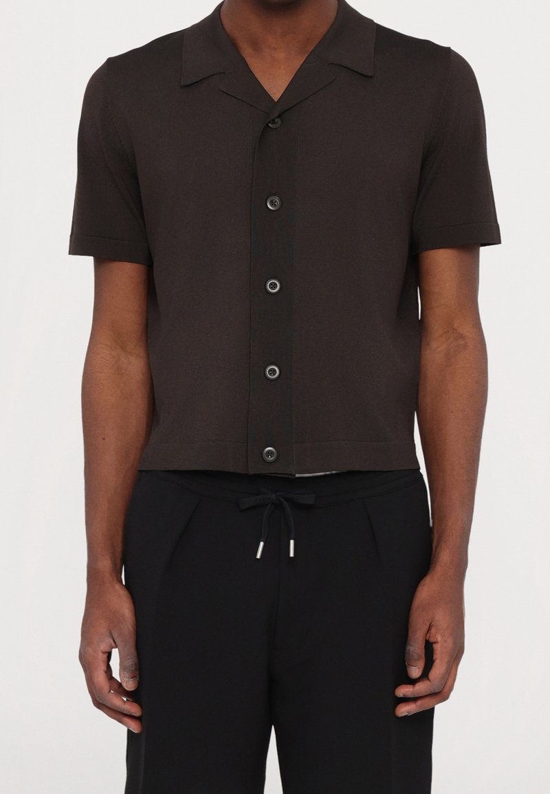 Homme portant une chemise noire à manches courtes avec col et un pantalon noir à cordon, debout devant un fond uni.