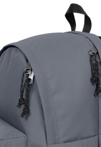 Mochila de tela gris con un diseño de parte superior redondeada, que presenta herrajes negros, cierres de cordón y una superficie texturizada. Incluye un asa superior resistente.