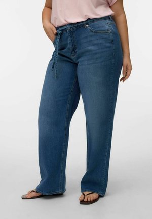 Personne portant un jean large bleu taille haute avec une ceinture en tissu, un haut rose clair et des sandales tongs marron, debout avec une main dans la poche.