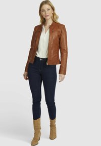 Chaqueta de cuero marrón con cremallera en el frente, mangas largas y dos bolsillos en el pecho con cremallera. Llevada sobre una camisa blanca y combinada con jeans oscuros y botas color beige.