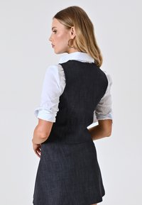 Gilet in denim scuro con un design aderente, abbinato a una camicia bianca con colletto. Presenta maniche arrotolate e una leggera texture, creando un look a strati.