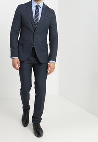 Abito blu marina con vestibilità su misura, giacca monopetto, rever a punta e pantaloni coordinati. Abbinato a una camicia azzurra chiara e cravatta a righe.
