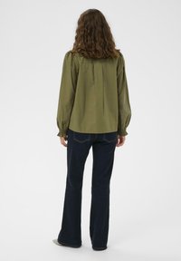 Olivno zelena bluzica s dolgimi rokavi, napihnjenimi rameni, nabranim hrbtom in nabranimi manšetami, v kombinaciji s temno modrimi flared jeans. Gladka tekstura.