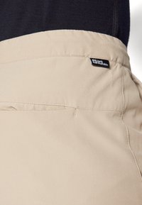 Bežové kalhoty s hladkým texturem, se zadní kapsou a černou značkovou etiketou na pase. Jednoduchý a funkční design.