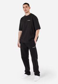 Zwart oversized T-shirt en bijpassende broek, beide van zachte stof. T-shirt heeft een wit logo; de broek heeft zijzakken en logo-details.