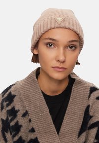 Berretto rosa lavorato a maglia con design a coste e un accento triangolare con logo dorato. Indossato con un cardigan a scialle con motivo sopra una maglietta nera.