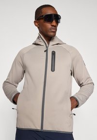 Beige zip-up takki, jossa on istuva huppu, pystysuora vetoketjupussi, harmaat yksityiskohdat ja elastiset hihansuut. Sileä, kevyt materiaali.
