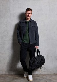 Szara wełniana kurtka na zielonej koszuli, czarne spodnie, białe trampki z zielonymi akcentami i czarna torba sportowa. Betonowe tło.