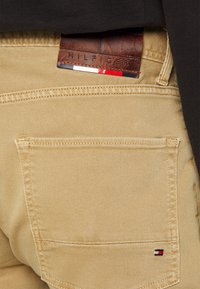 Beige puuvillachinot, joissa on minimaalinen venyvyys. Vyötäröllä on nahkaetiketti, johon on kirjailtu "HILFIGER". Mukana pieni logoitse.