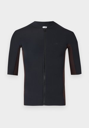Mann som står barfot, iført tettsittende sort kortermet genser med glidelås og matchende knelengde shorts mot en ensfarget hvit bakgrunn.