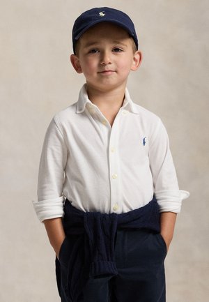 Chemise blanche à manches longues avec fermeture à boutons, cardigan navy noué à la taille, et casquette navy avec logo. Enfant debout les mains dans les poches.