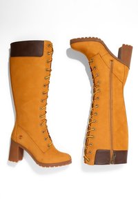 Botas altas con cordones en ante amarillo mostaza y parte superior de cuero marrón, que cuentan con un tacón grueso y cremallera lateral para el cierre.