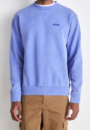 Man draagt een lichtpaarse trui met ronde hals met het "BOSS"-logo en bruine cargobroek, staand tegen een effen lichte achtergrond.