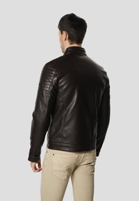 Pruun nahkjakk tikitud õlakatete, seiseva krae ja liibuva lõikega, kombineeritud beežide pükstega. Tagantvaade.