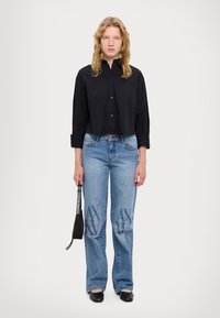 Camicia nera corta con bottoni, jeans azzurri a palazzo con accentuazione del logo. Una borsa nera e delle scarpe slip-on completano il look.