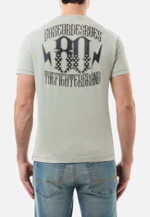 Mann trägt ein hellgraues T-Shirt mit der Aufschrift „Boxeur Des Rues 80 The Fighter Brand“ und Blitzgrafik auf dem Rücken, kombiniert mit blauen Jeans.