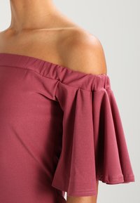 Haut à épaules dénudées dans une teinte mauve unie, avec des manches à volants à design élastiqué froncé, texture douce et silhouette ajustée.